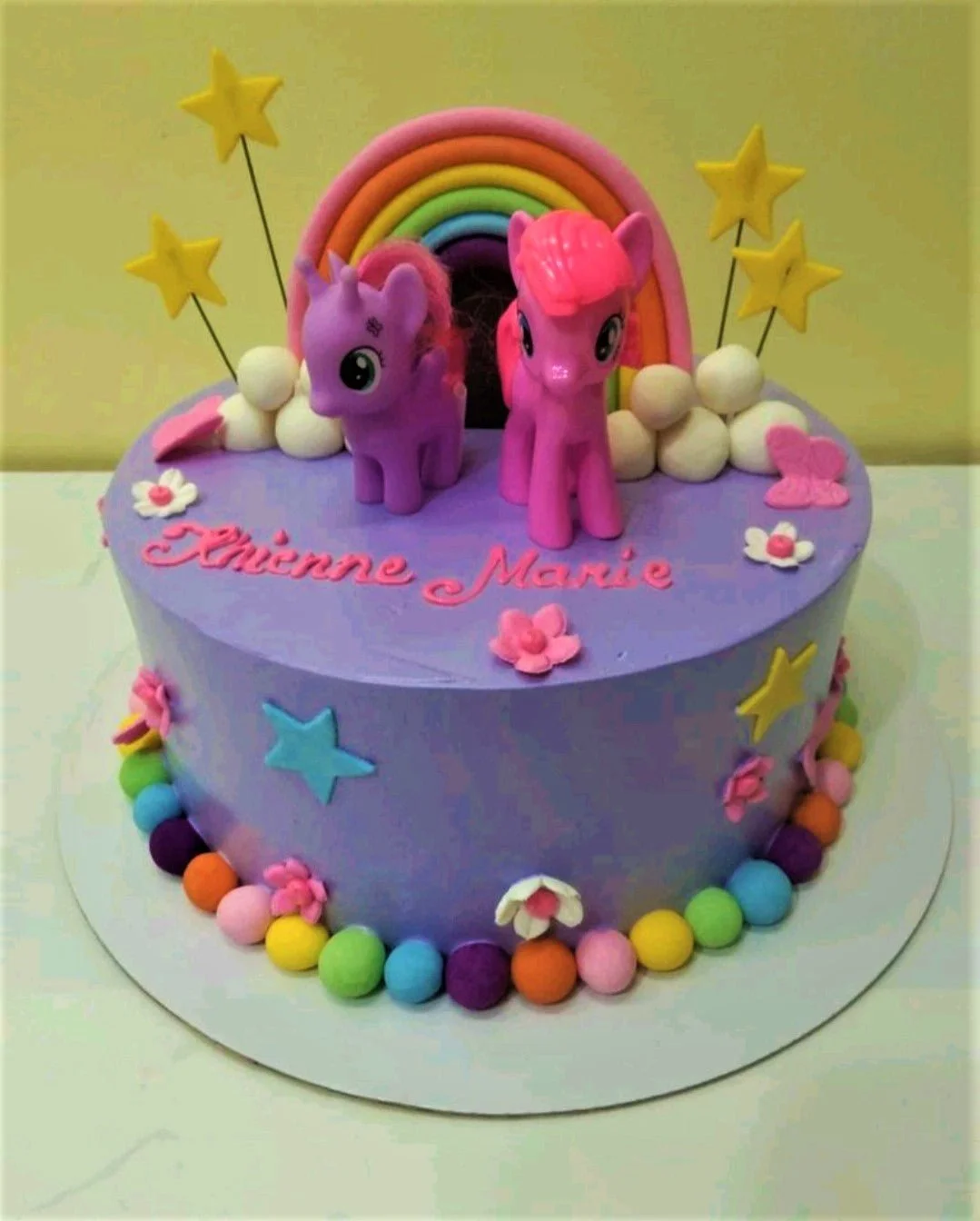 My Little Pony-Inspired Sky Blue Birthday Cake A purple 1 Tier cake themed around My Little Pony, featuring two 3D pony figures, a rainbow, and colorful fondant balls.