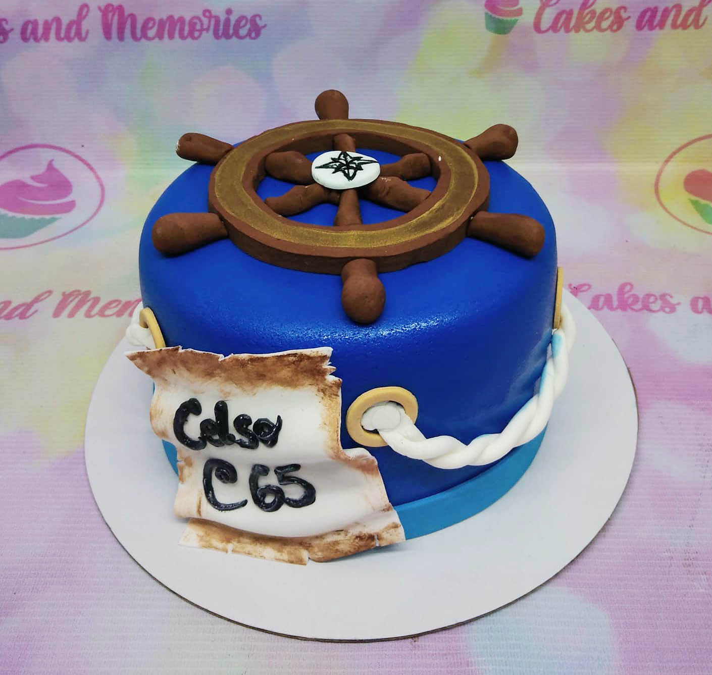 Blue nautical themed cake with a large brown ship wheel topper and a fondant scroll message on the side. Blue nautical themed cake with a large brown ship wheel topper and a fondant scroll message on the side.