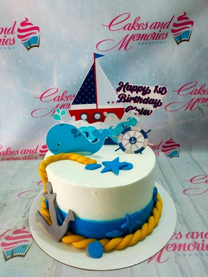 Nautical Birthday Cake A white, blue, and yellow 1-tier cake themed Nautical, featuring a sailboat, whale, ship's wheel, and anchor.