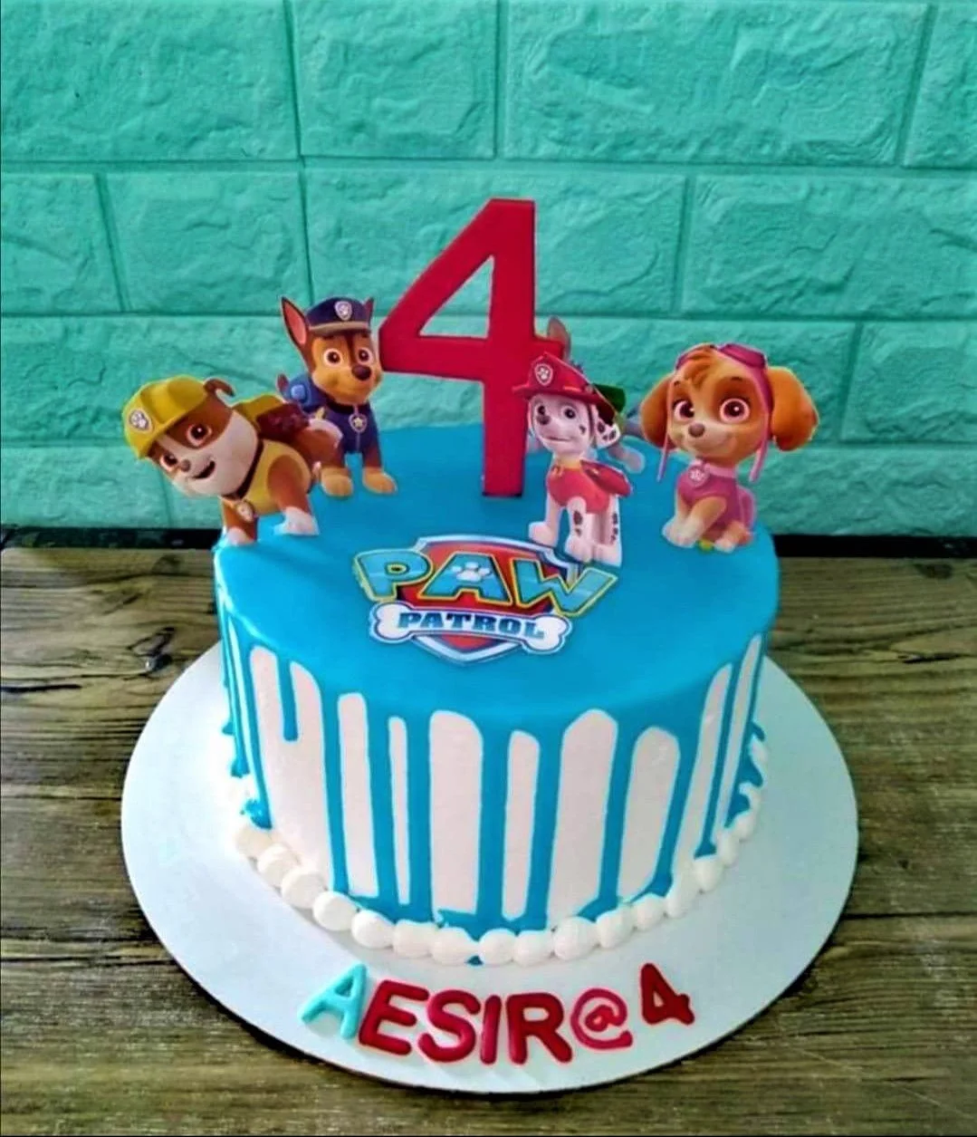 A blue and white 1-tier Paw Patrol cake, with character printouts and a red number 4 topper. A blue and white 1-tier Paw Patrol cake, with character printouts and a red number 4 topper.