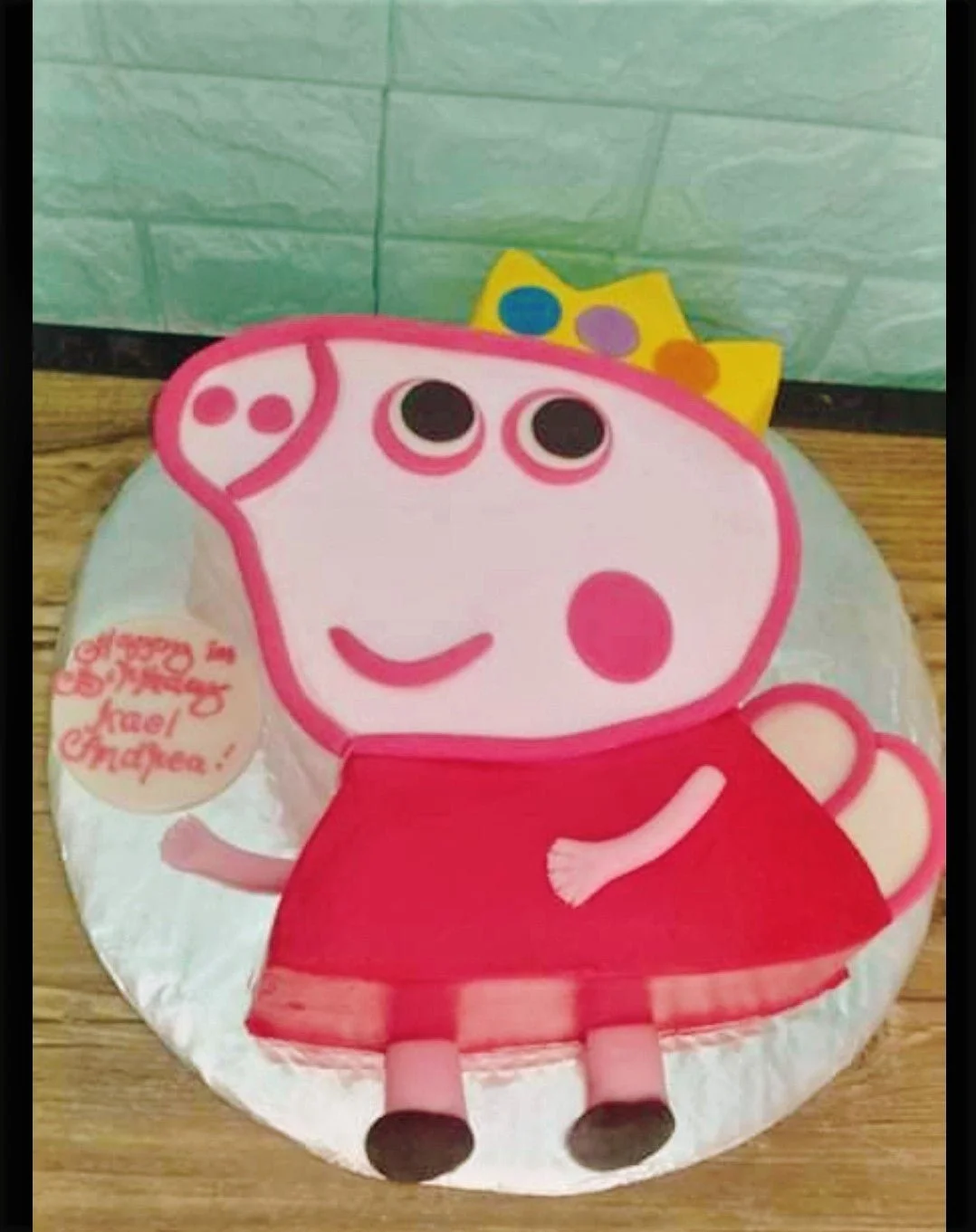 Peppa Pig shaped fondant cake with a yellow crown and a pink dress on a silver board.