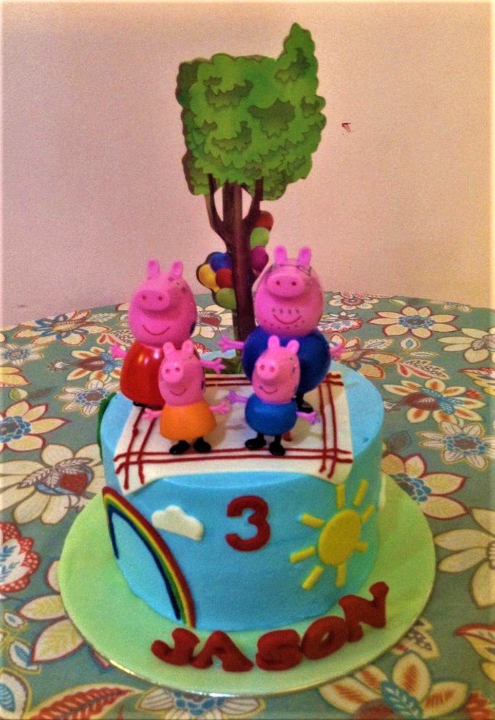 Light blue Peppa Pig themed birthday cake with 3D character figures, a large tree printout, and a fondant rainbow and sun on the side. Light blue Peppa Pig themed birthday cake with 3D character figures, a large tree printout, and a fondant rainbow and sun on the side.