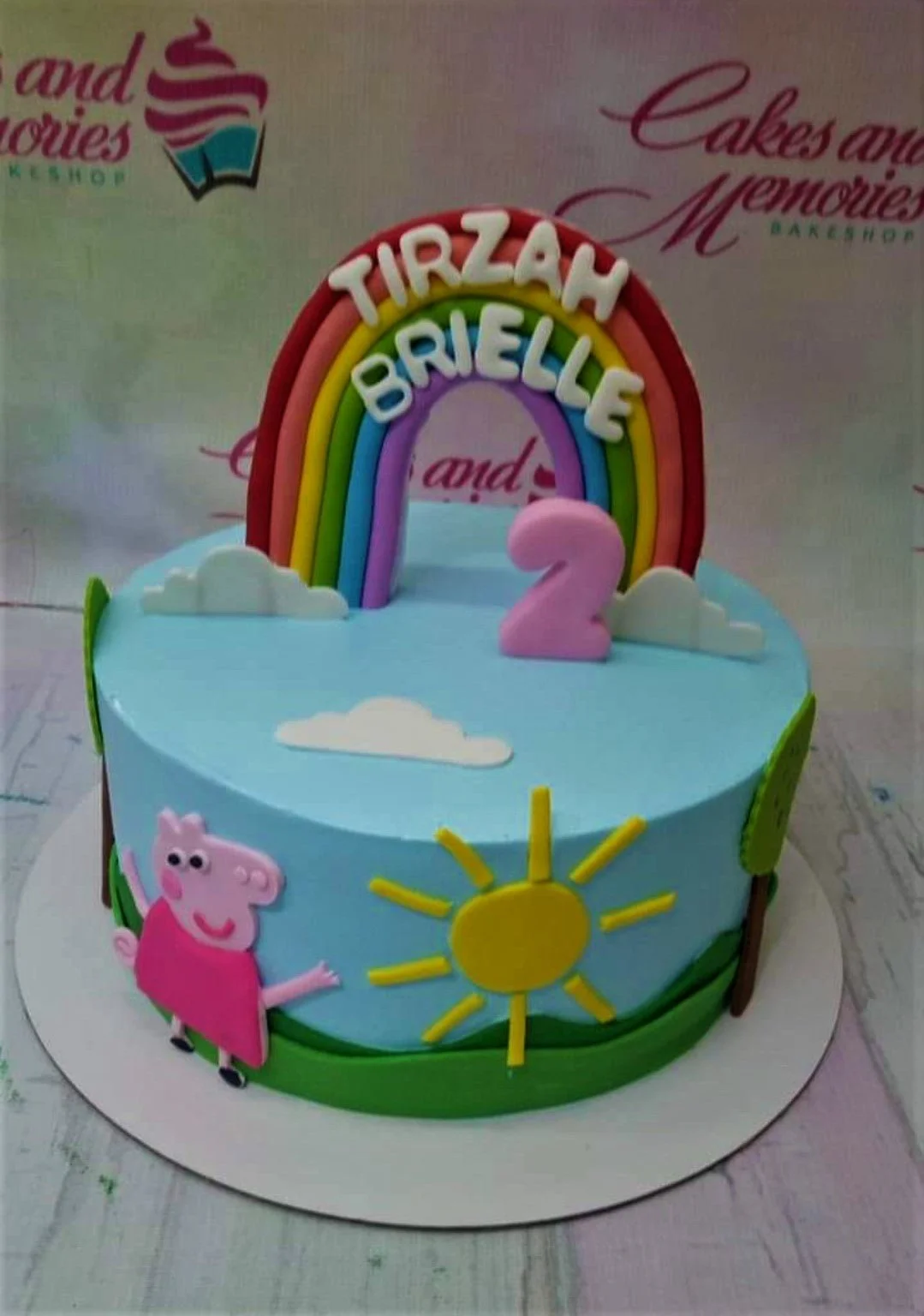 Light blue Peppa Pig themed birthday cake with a large fondant rainbow, pink number 2, and Peppa Pig character on the side.
