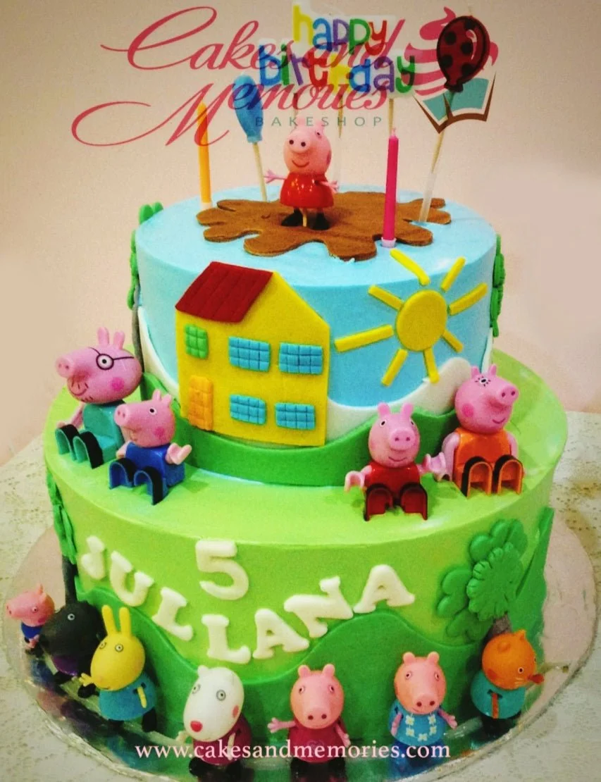 Two-tier Peppa Pig themed birthday cake with green and blue fondant, featuring various character figurines and a yellow house decoration.
