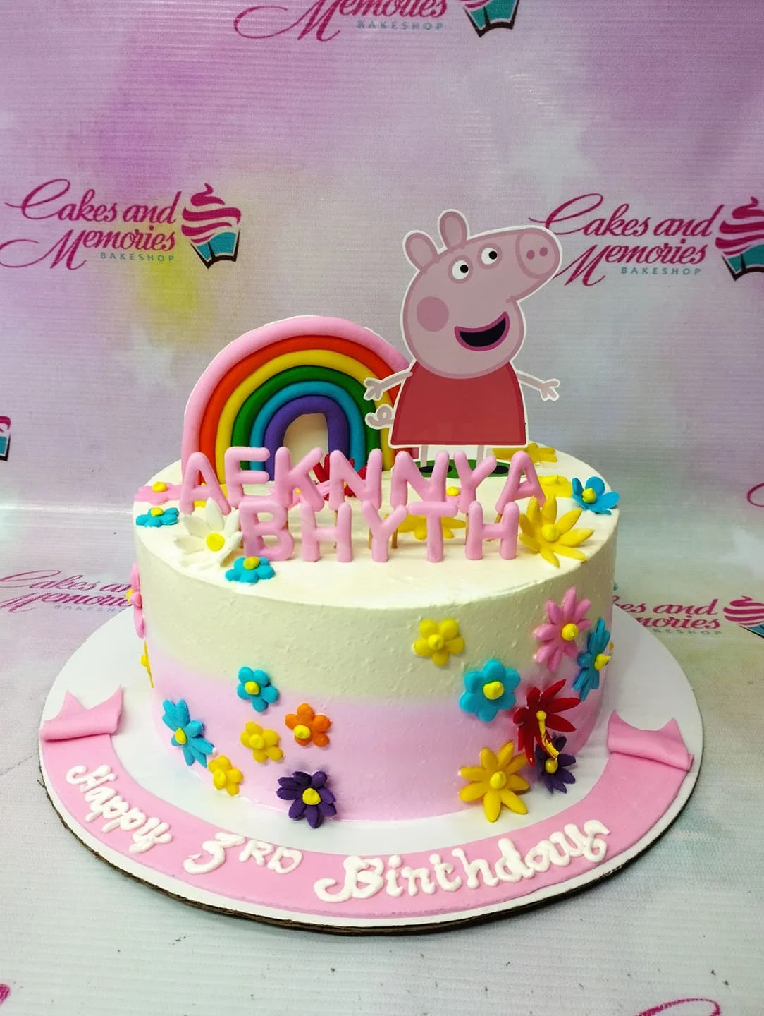 Peppa Pig themed birthday cake with a rainbow topper, colorful fondant flowers, and a pink message banner on the base.