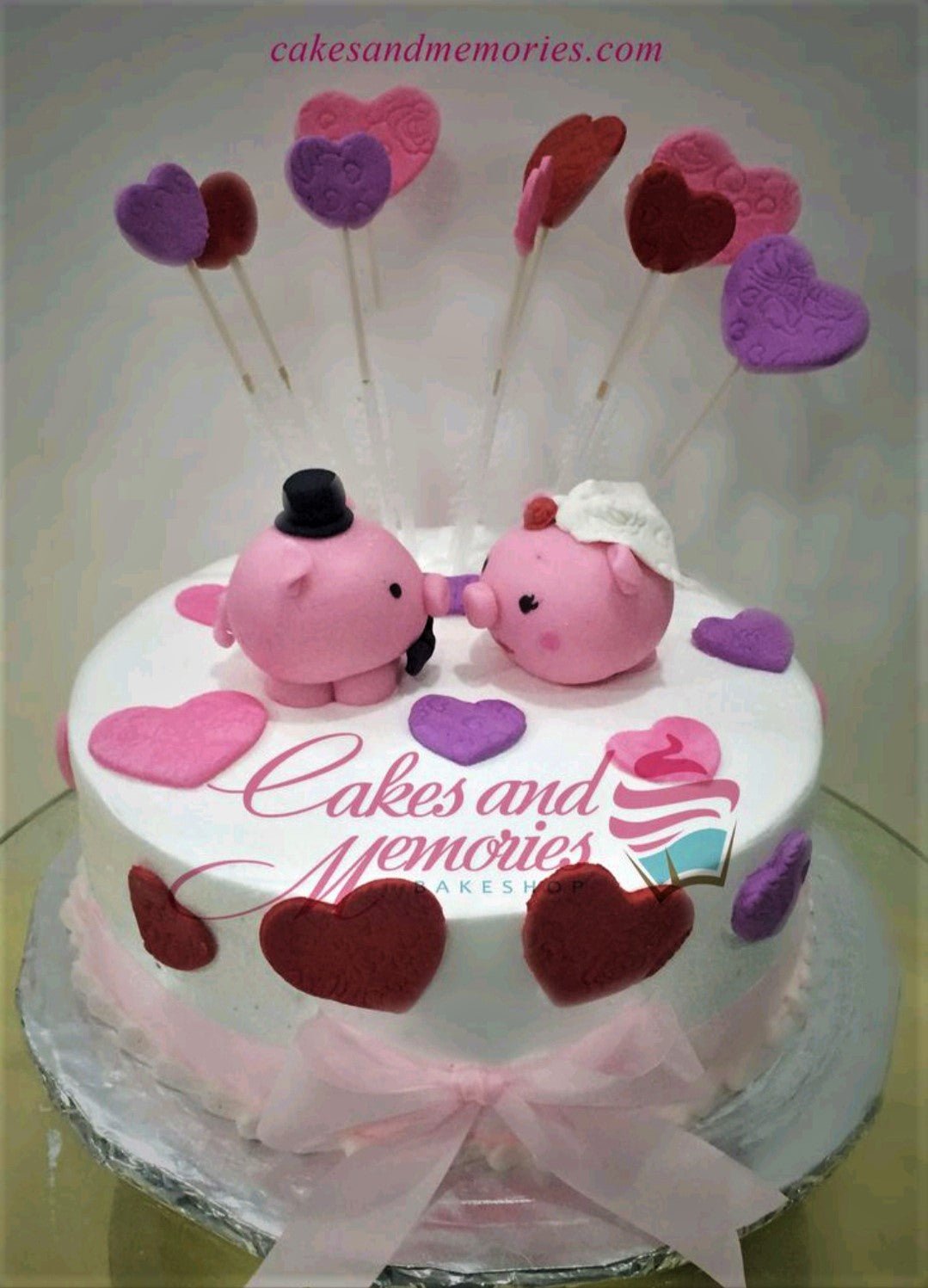 White round cake with pink pig bride and groom fondant toppers, pink and purple hearts, and a pink ribbon bow. White round cake with pink pig bride and groom fondant toppers, pink and purple hearts, and a pink ribbon bow.