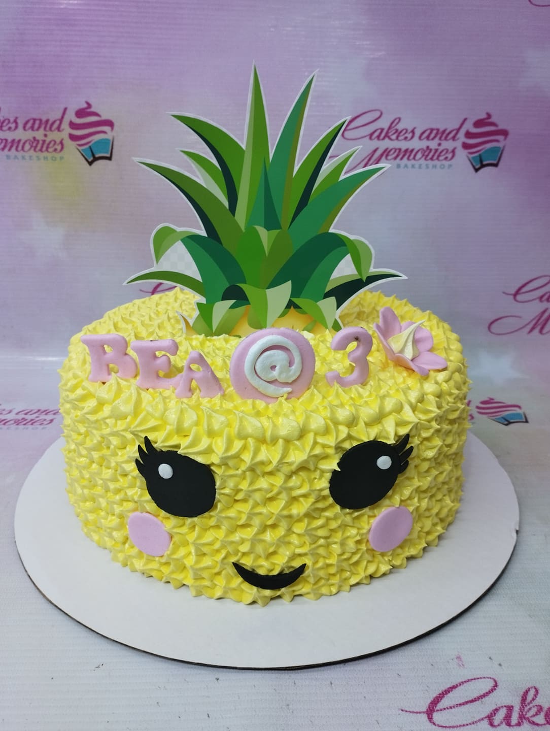 Yellow pineapple-themed birthday cake with piped icing texture, pink fondant letters spelling BEA @ 3, and a large green pineapple leaf topper. Yellow pineapple-themed birthday cake with piped icing texture, pink fondant letters spelling BEA @ 3, and a large green pineapple leaf topper.