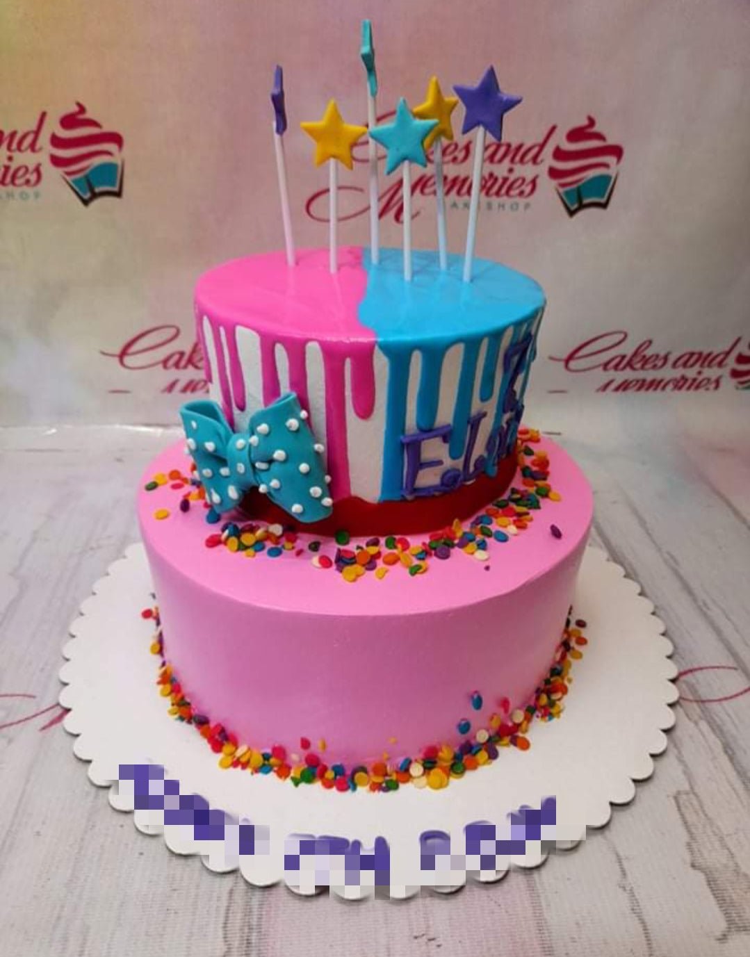 Two-tier pink and blue drip cake with colorful sprinkles, a blue polka dot bow, and star toppers on sticks. Two-tier pink and blue drip cake with colorful sprinkles, a blue polka dot bow, and star toppers on sticks.