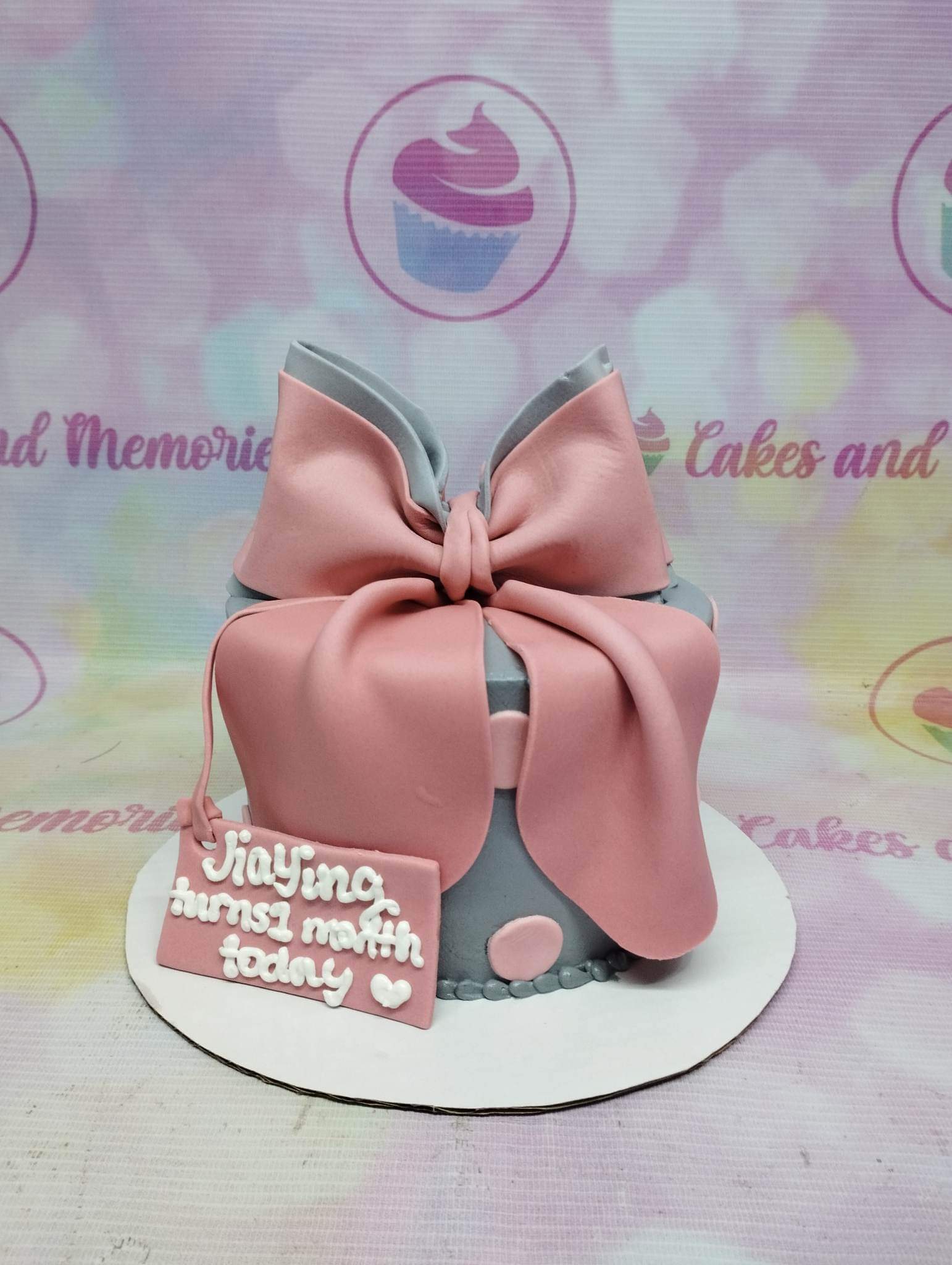 Gray fondant cake with a large pink fondant bow and a personalized message tag for a one-month celebration. Gray fondant cake with a large pink fondant bow and a personalized message tag for a one-month celebration.