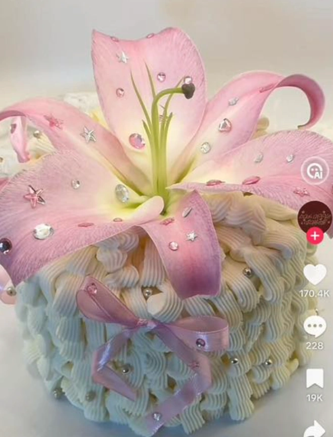 White soft-icing cake with intricate piped ruffles, a large pink lily topper adorned with gems, and a pink ribbon bow. White soft-icing cake with intricate piped ruffles, a large pink lily topper adorned with gems, and a pink ribbon bow.