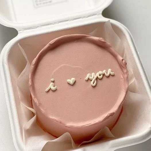 Minimalist pink bento cake with white piped icing message 'i heart you' in a white takeout container. Minimalist pink bento cake with white piped icing message 'i heart you' in a white takeout container.