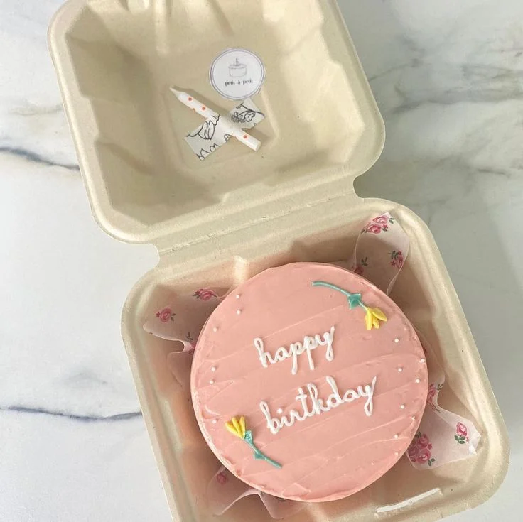 Pink bento cake with white piped Happy Birthday message and yellow icing flowers in a compostable box with a candle. Pink bento cake with white piped Happy Birthday message and yellow icing flowers in a compostable box with a candle.