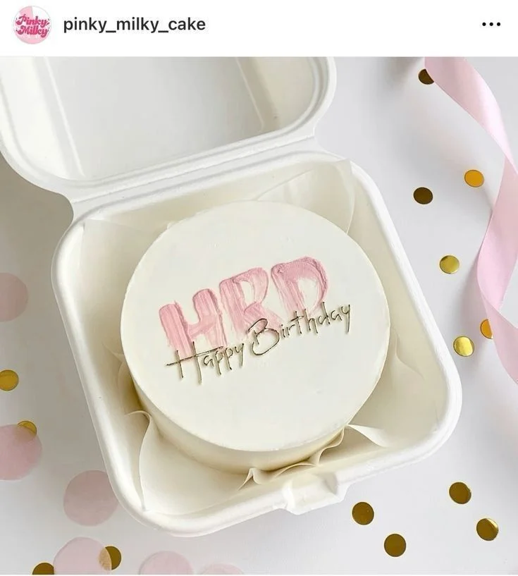 White bento cake with pink palette knife 'HBD' text and a gold 'Happy Birthday' cardstock topper in a white container. White bento cake with pink palette knife 'HBD' text and a gold 'Happy Birthday' cardstock topper in a white container.