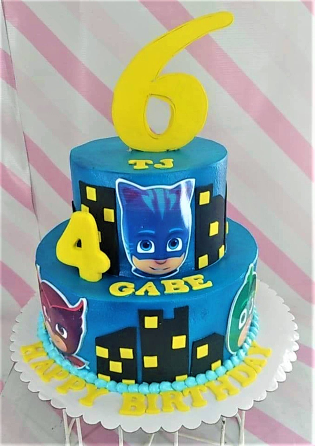 Two-tier blue PJ Masks themed birthday cake with character printouts, yellow fondant numbers, and a city skyline design. Two-tier blue PJ Masks themed birthday cake with character printouts, yellow fondant numbers, and a city skyline design.