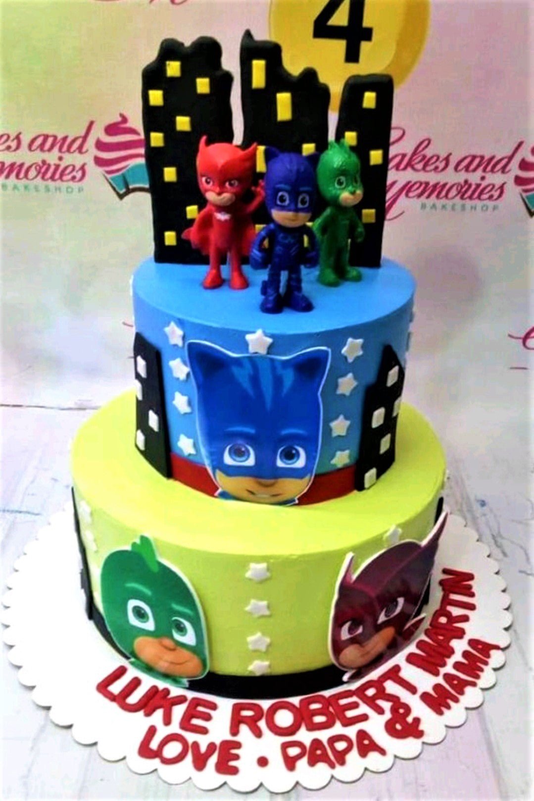 Two-tier PJ Masks themed cake with blue and lime green icing, featuring Catboy, Owlette, and Gekko figurines and city skyline toppers. Two-tier PJ Masks themed cake with blue and lime green icing, featuring Catboy, Owlette, and Gekko figurines and city skyline toppers.