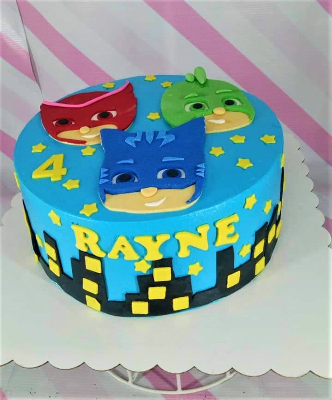 Blue PJ Masks themed birthday cake with Catboy, Owlette, and Gekko fondant faces, yellow stars, and a city skyline border. Blue PJ Masks themed birthday cake with Catboy, Owlette, and Gekko fondant faces, yellow stars, and a city skyline border.