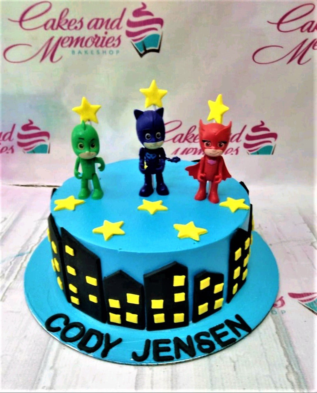 Blue PJ Masks themed birthday cake with Gekko, Catboy, and Owlette toy figurines and black fondant city skyline. Blue PJ Masks themed birthday cake with Gekko, Catboy, and Owlette toy figurines and black fondant city skyline.
