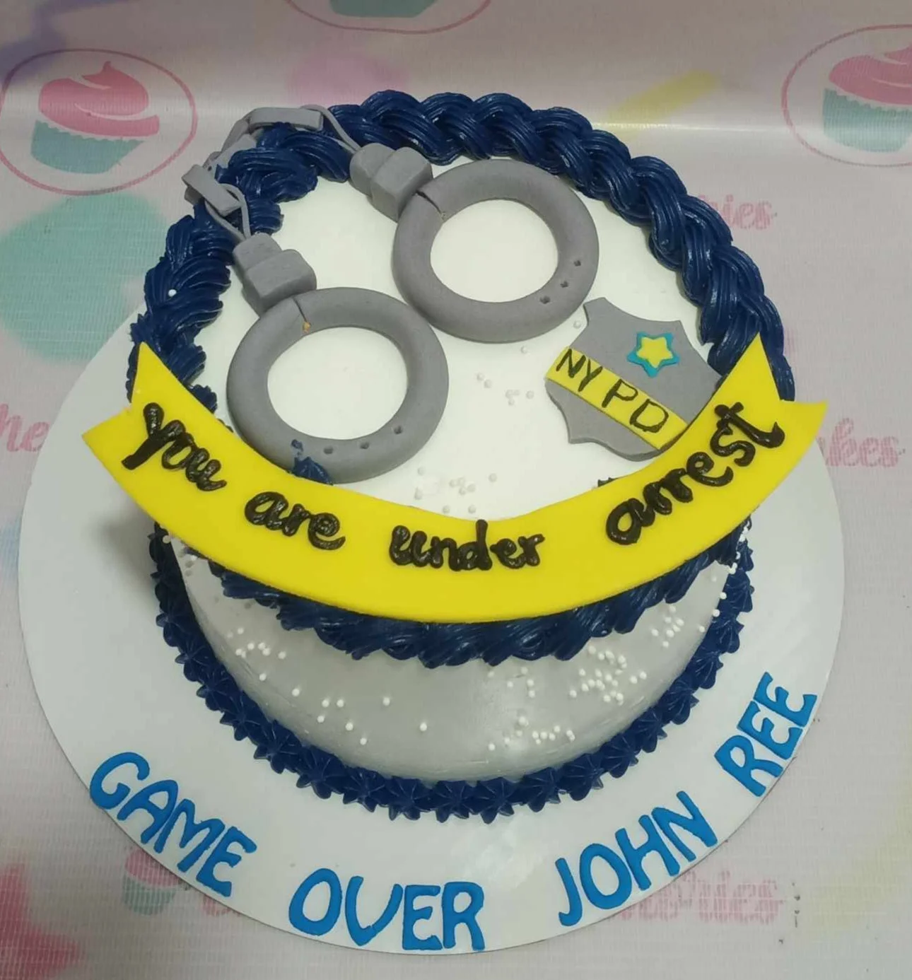 Police themed birthday cake with grey fondant handcuffs, NYPD badge, and yellow banner on white icing with navy blue borders. Police themed birthday cake with grey fondant handcuffs, NYPD badge, and yellow banner on white icing with navy blue borders.