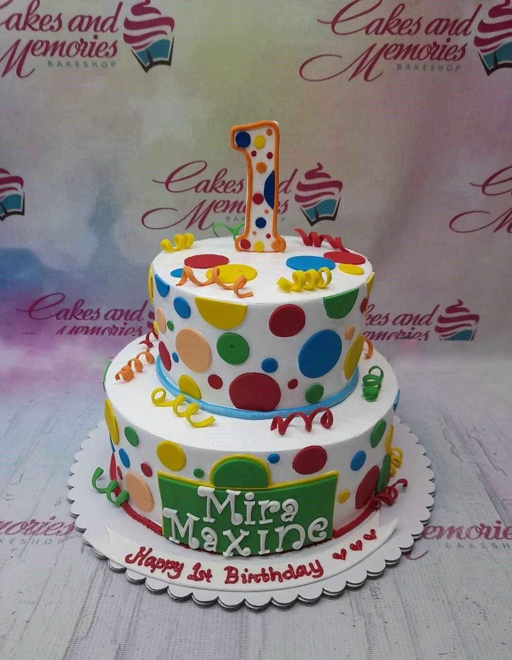Polka Dot Birthday White 2-Tier Cake A white and colorful 2-tier Polka Dot Birthday cake, featuring a large number 1 topper, multi-colored polka dots, and a green name plaque.