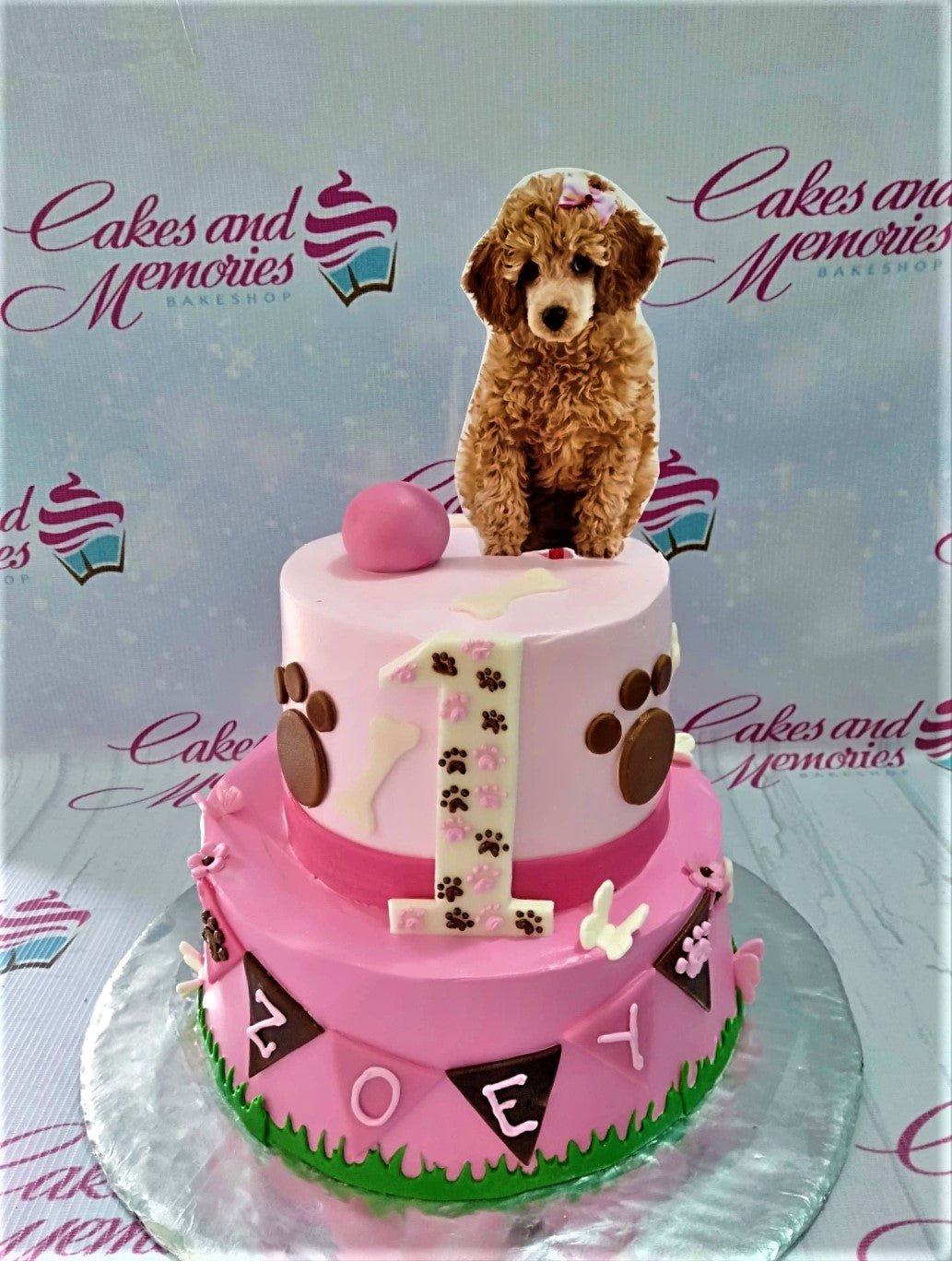 Two-tier pink poodle themed birthday cake with a large poodle printout topper, paw prints, and a number one candle. Two-tier pink poodle themed birthday cake with a large poodle printout topper, paw prints, and a number one candle.