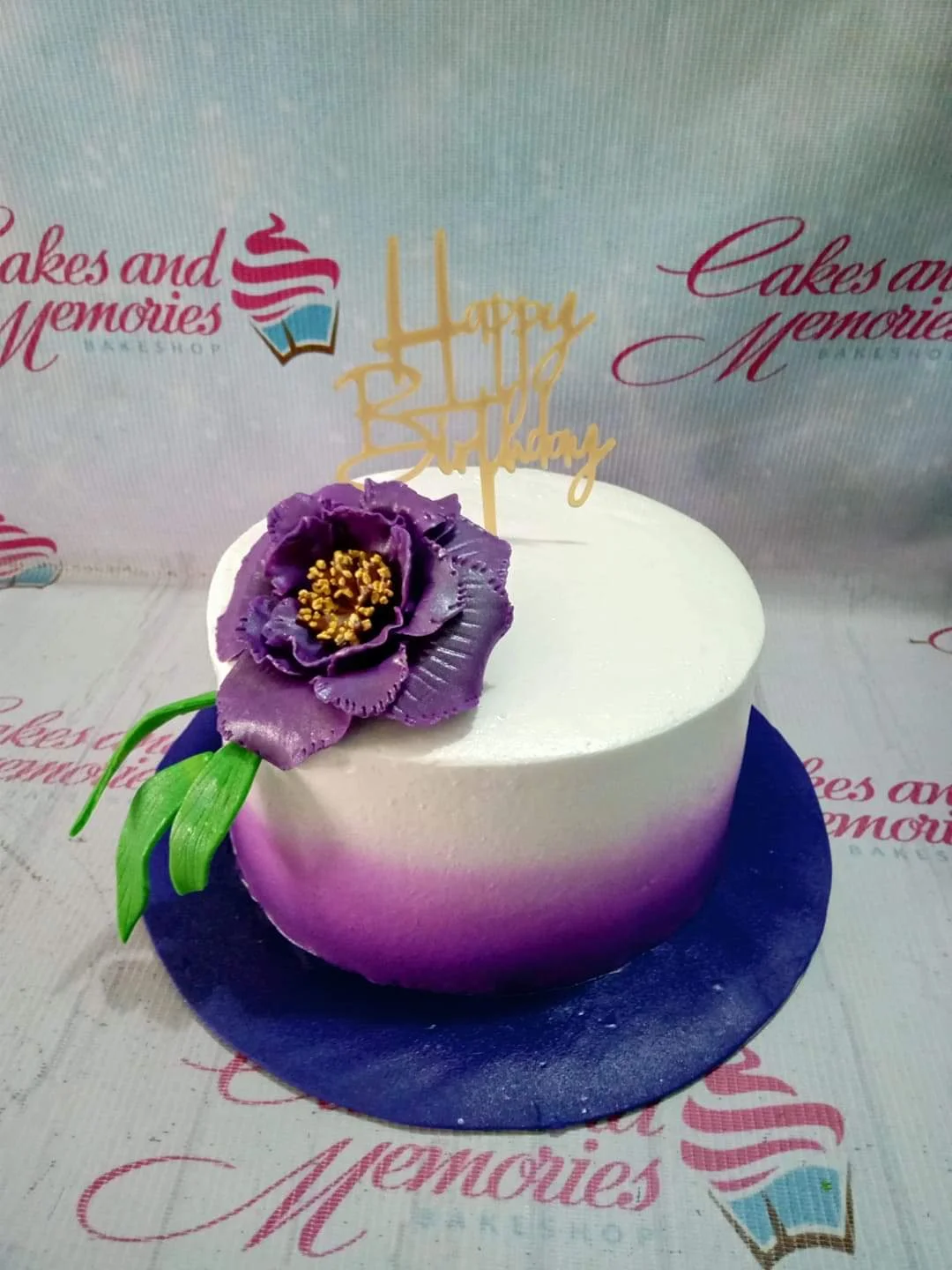 Purple Floral White Birthday Cake A white and purple gradient 1 Tier cake, floral themed, with a large purple gumpaste flower, a gold 'Happy Birthday' topper, and green leaves.