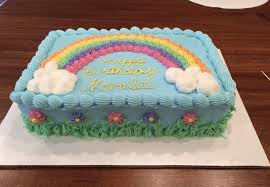 Rectangular light blue birthday cake with a piped rainbow, white clouds, and colorful flowers along a green grass border. Rectangular light blue birthday cake with a piped rainbow, white clouds, and colorful flowers along a green grass border.
