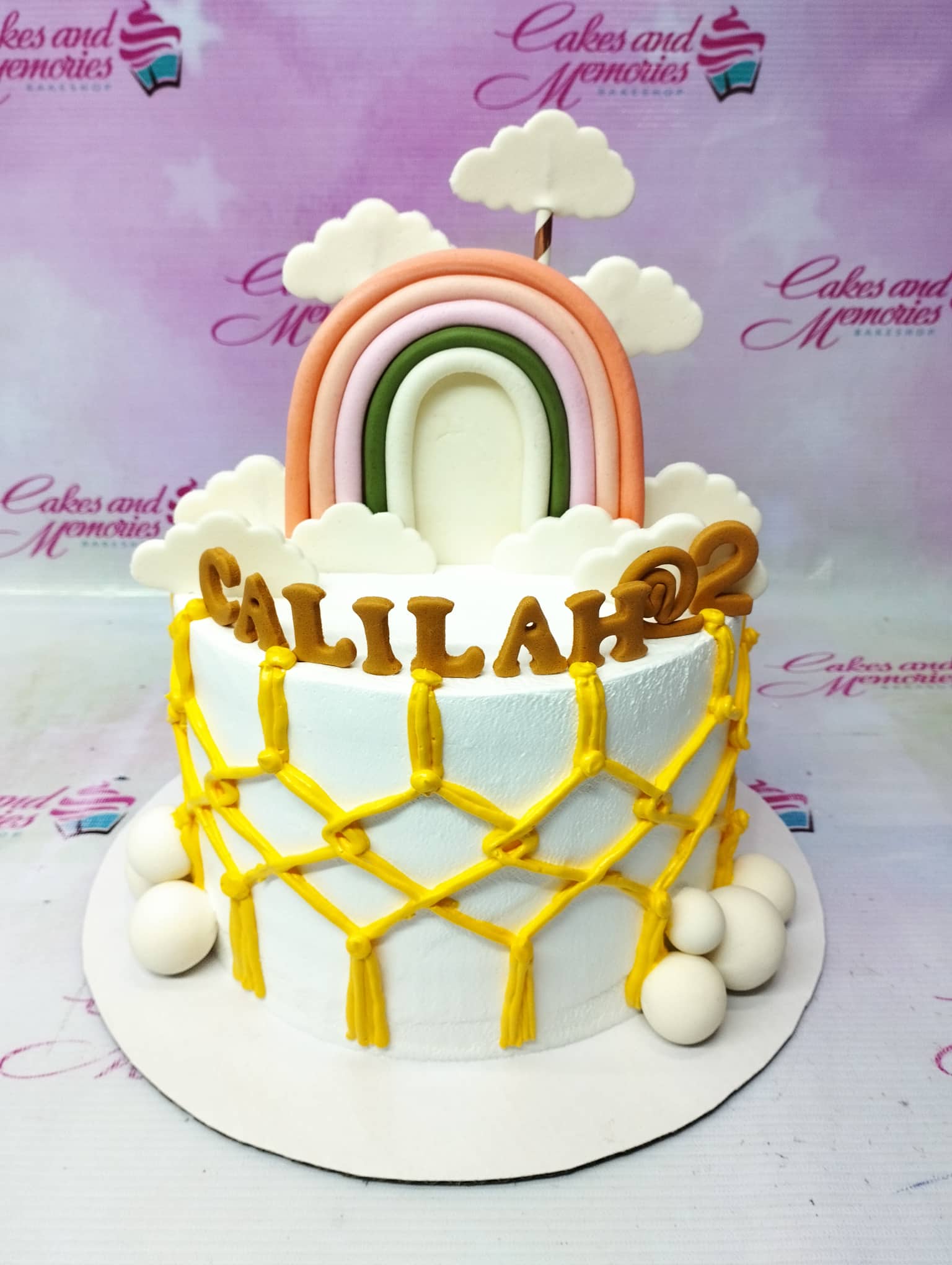 White 1-tier birthday cake with a large fondant rainbow, white clouds, and yellow macrame-style icing design. White 1-tier birthday cake with a large fondant rainbow, white clouds, and yellow macrame-style icing design.