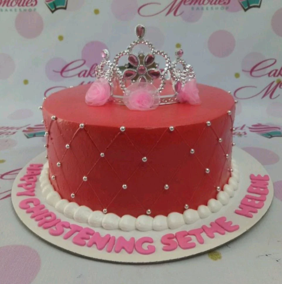Red quilted cake with silver dragees, a silver rhinestone tiara, and pink fabric flowers on a white base board with pink lettering.
