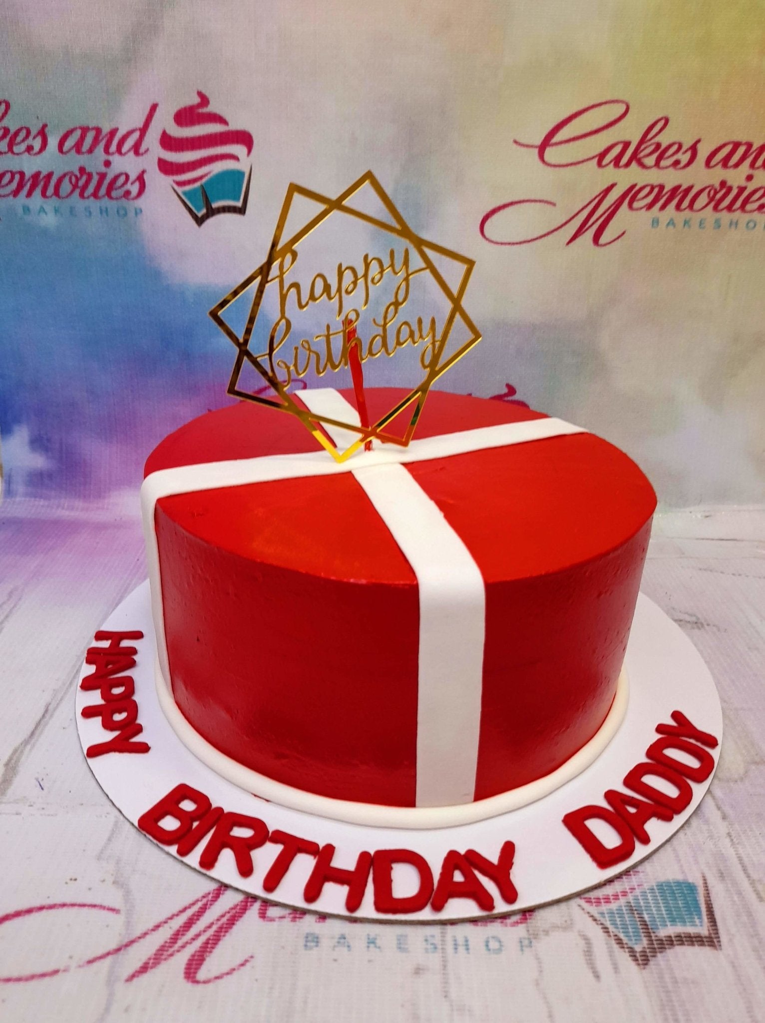 Red minimalist gift box cake with white fondant ribbons and a gold Happy Birthday topper on a white base board with red lettering.