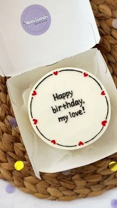 White minimalist bento cake with black piped icing text and small red heart decorations around the top edge. White minimalist bento cake with black piped icing text and small red heart decorations around the top edge.