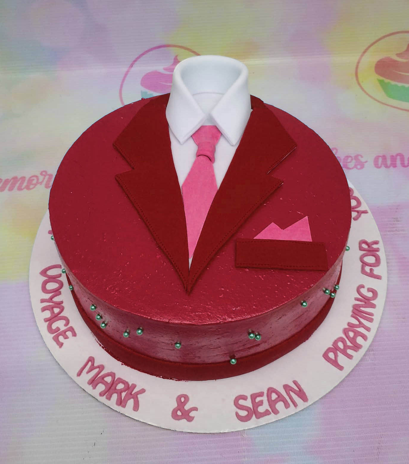 Red tuxedo suit cake with a white shirt and pink tie, featuring green pearl accents and a personalized message on the base board. Red tuxedo suit cake with a white shirt and pink tie, featuring green pearl accents and a personalized message on the base board.