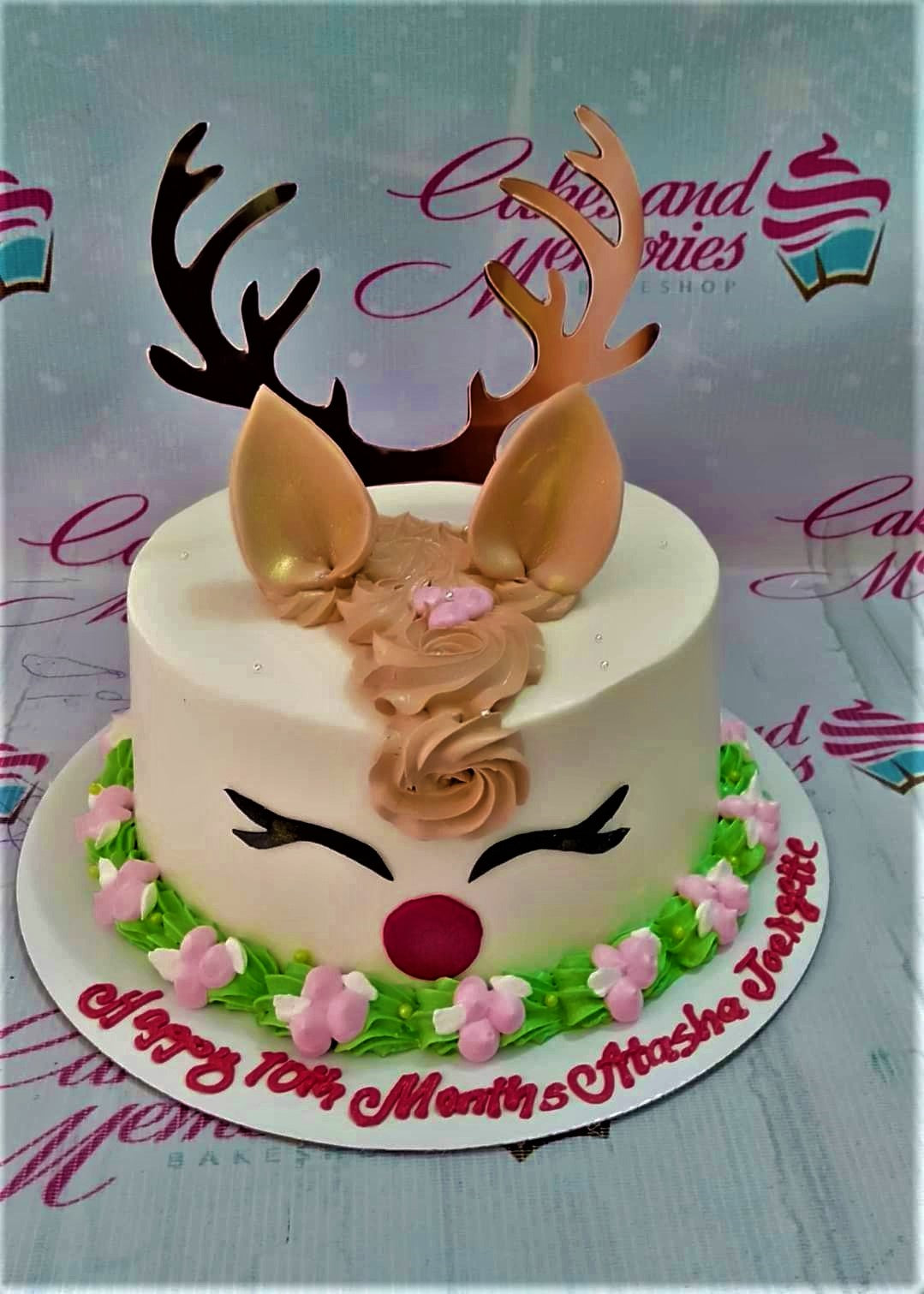 White reindeer cake with gold antlers, brown icing mane, and pink floral border on a white base board. White reindeer cake with gold antlers, brown icing mane, and pink floral border on a white base board.