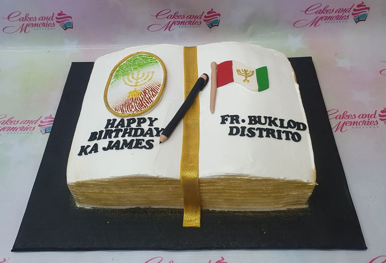 Open book birthday cake with white icing, gold ribbon bookmark, and religious symbols for Ka James. Open book birthday cake with white icing, gold ribbon bookmark, and religious symbols for Ka James.
