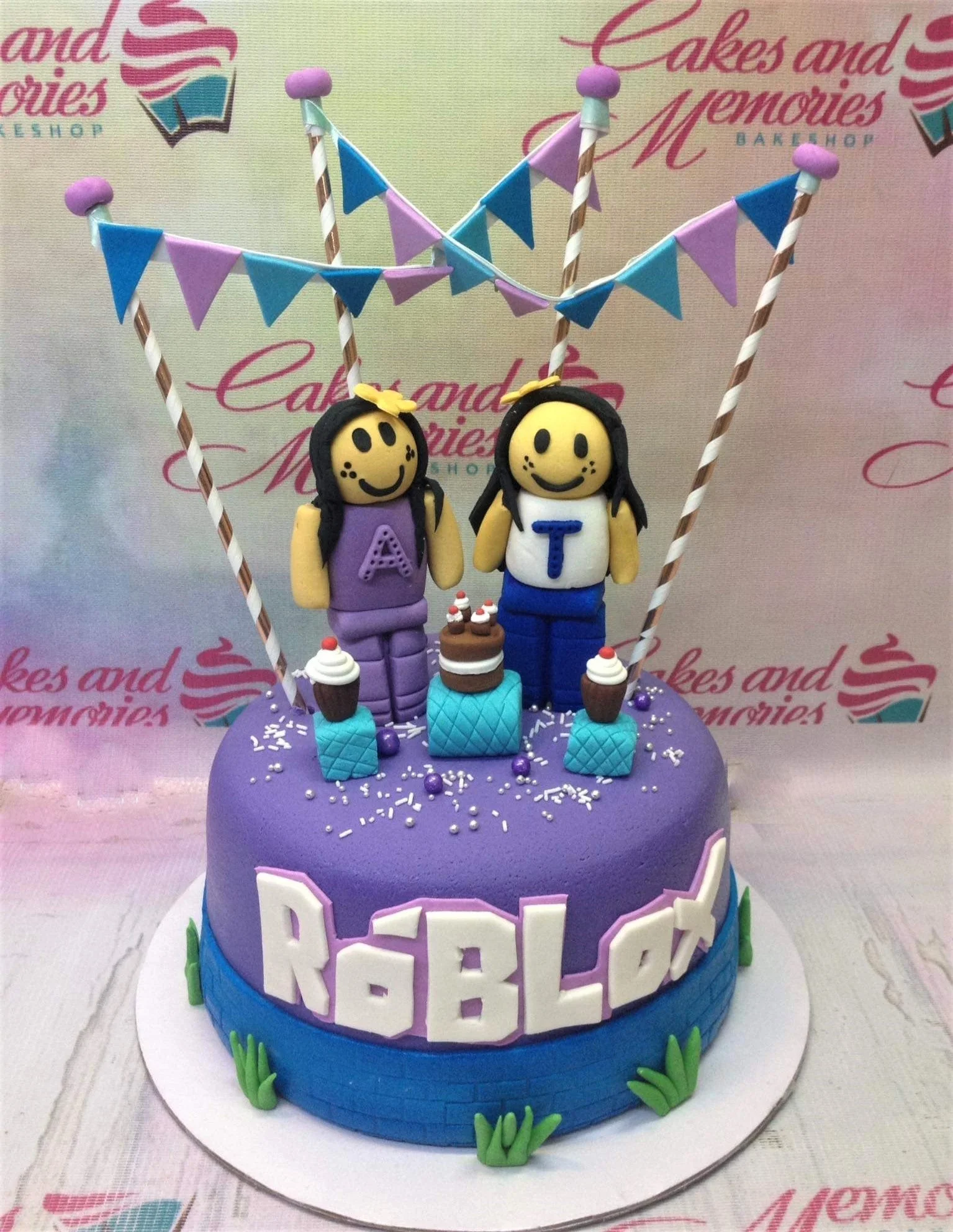 Roblox-Inspired Cake A purple and blue fondant cake themed around Roblox, featuring two girl figures, bunting flags, and a brick-textured base.