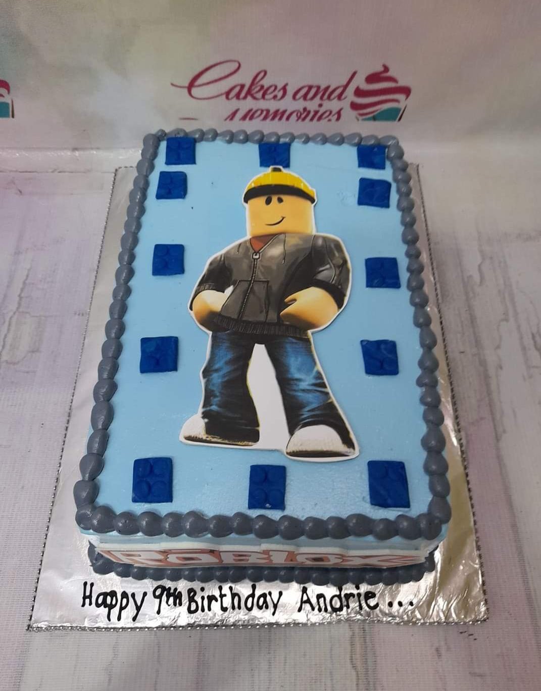 Light blue rectangular Roblox themed birthday cake with a large character printout and dark gray icing borders. Light blue rectangular Roblox themed birthday cake with a large character printout and dark gray icing borders.