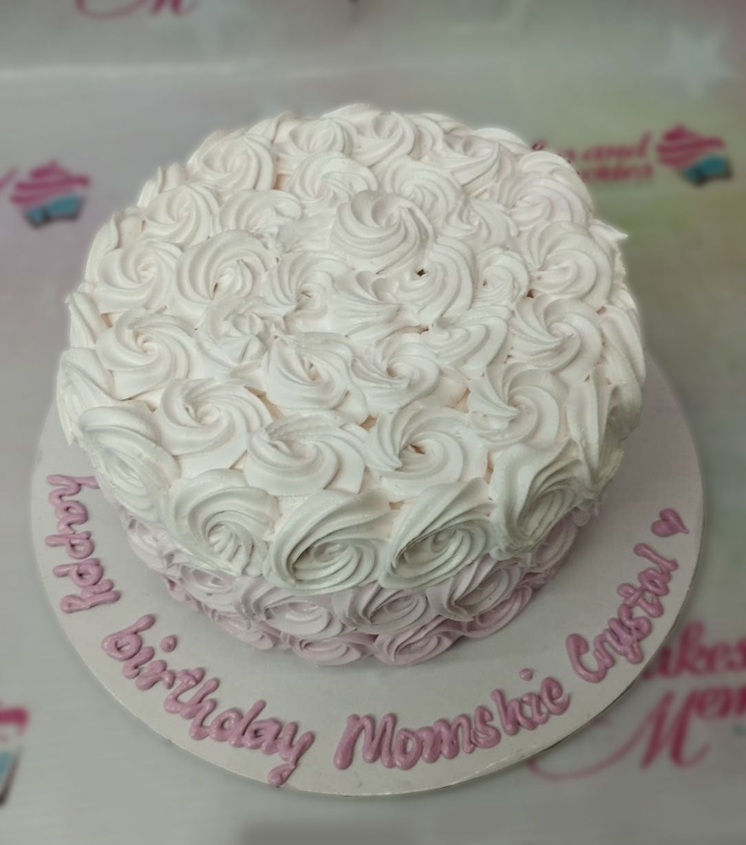 A single-tier round cake covered in white and light pink icing rosettes with a birthday message on the base board. A single-tier round cake covered in white and light pink icing rosettes with a birthday message on the base board.