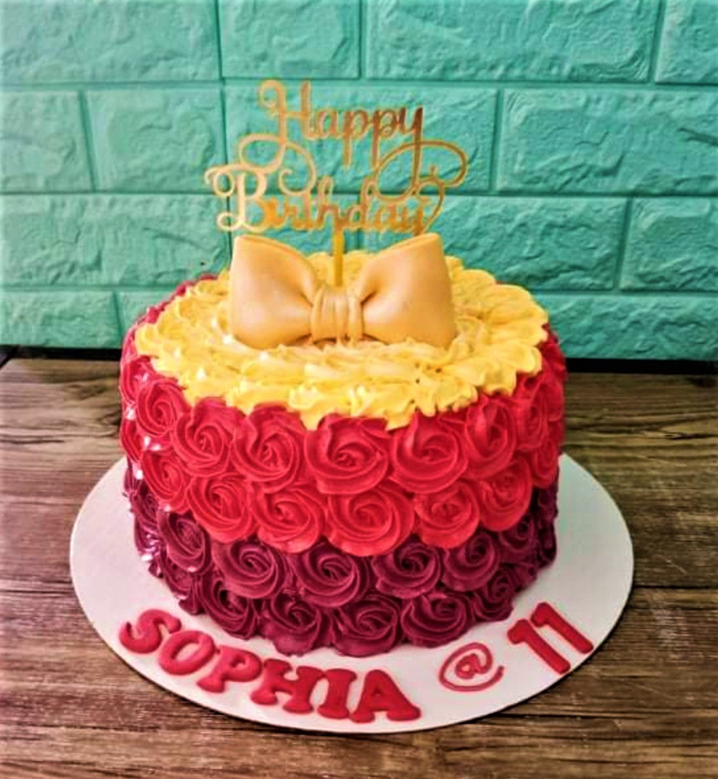 A round birthday cake with red and purple icing rosettes, a large gold fondant bow, and a gold Happy Birthday topper. A round birthday cake with red and purple icing rosettes, a large gold fondant bow, and a gold Happy Birthday topper.