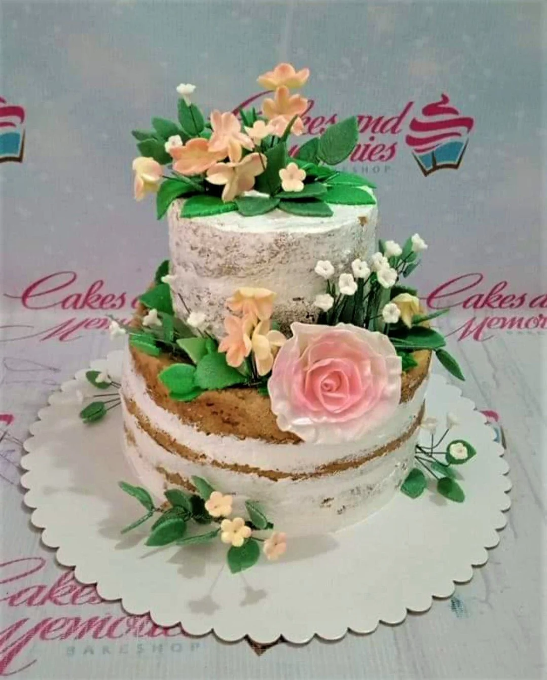 Two-tier semi-naked rustic cake decorated with peach and pink edible flowers and green leaves on a white scalloped base.