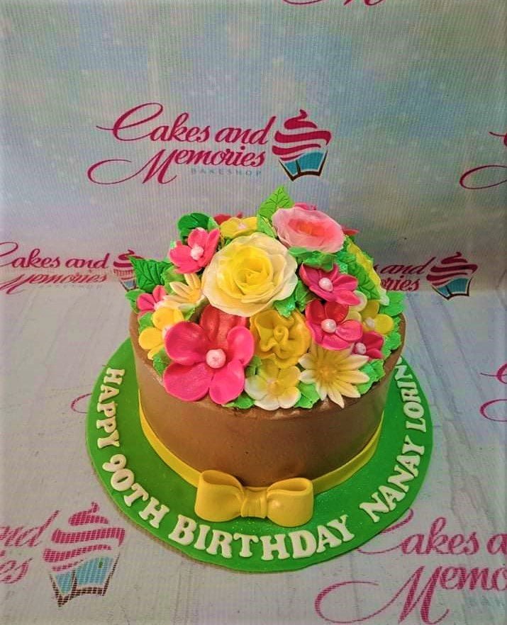 Chocolate brown round cake topped with a dense bouquet of pink, yellow, and white edible flowers with green leaves and a yellow fondant bow. Chocolate brown round cake topped with a dense bouquet of pink, yellow, and white edible flowers with green leaves and a yellow fondant bow.