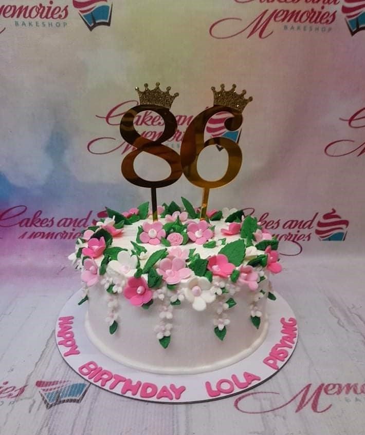 White 1-tier birthday cake decorated with pink and white fondant flowers and gold number 86 toppers with crowns. White 1-tier birthday cake decorated with pink and white fondant flowers and gold number 86 toppers with crowns.