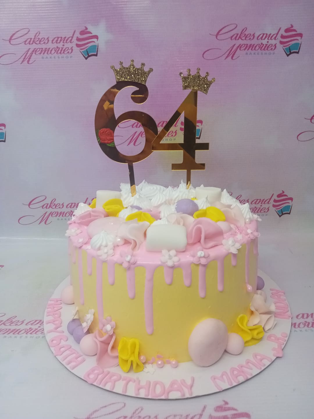 Yellow 1-tier birthday cake with pink drip, white icing swirls, marshmallows, and gold number 64 toppers with crowns. Yellow 1-tier birthday cake with pink drip, white icing swirls, marshmallows, and gold number 64 toppers with crowns.