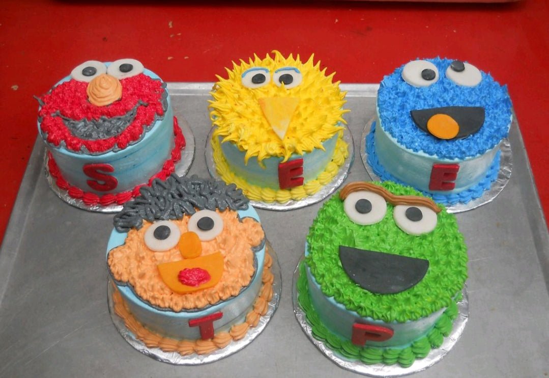 Five individual mini cakes featuring Sesame Street characters Elmo, Big Bird, Cookie Monster, Ernie, and Oscar the Grouch with piped icing. Five individual mini cakes featuring Sesame Street characters Elmo, Big Bird, Cookie Monster, Ernie, and Oscar the Grouch with piped icing.