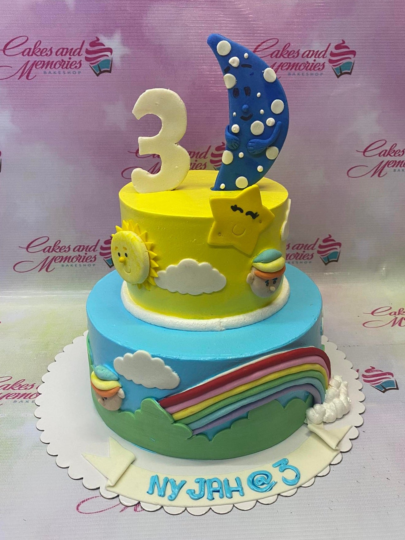 Two-tier sky-themed birthday cake with a yellow top tier and blue bottom tier featuring a fondant moon, sun, stars, and rainbow. Two-tier sky-themed birthday cake with a yellow top tier and blue bottom tier featuring a fondant moon, sun, stars, and rainbow.
