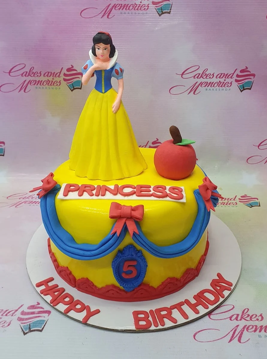 Snow White themed yellow fondant cake with a Snow White figurine, red apple topper, and blue icing drapes with red bows.