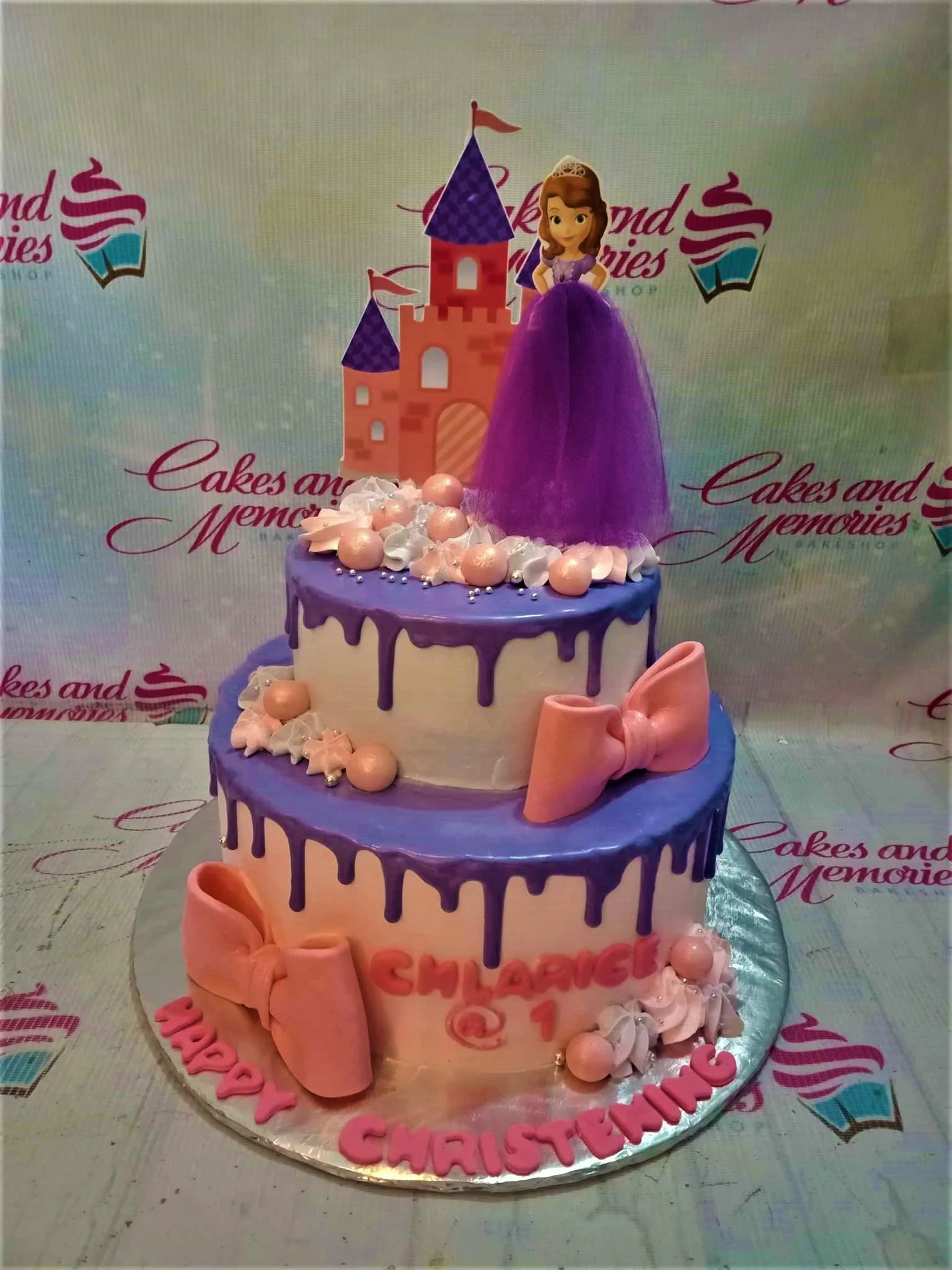Two-tier Sofia the First themed cake with purple drip icing, pink fondant bows, and a princess castle topper.