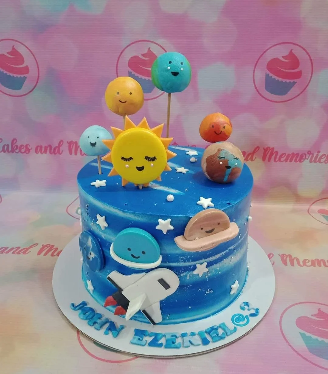 Space Sky Blue Birthday Cake A blue 1 Tier cake themed around Space, featuring a rocket ship, astronaut, sun, and planets, with star sprinkles and 'JOHN EZEKIEL @ 3' text.