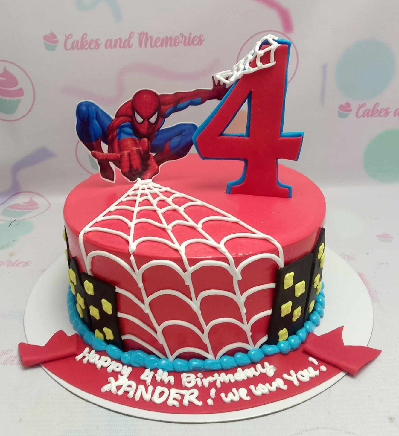 A red 1 Tier cake featuring a Spiderman character printout, a number 4 web topper, white spider web piping, black city skyline panels, and a blue pearl icing border. A red 1 Tier cake featuring a Spiderman character printout, a number 4 web topper, white spider web piping, black city skyline panels, and a blue pearl icing border.