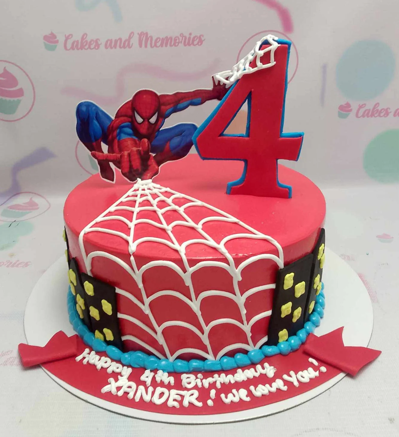 A red 1 Tier cake featuring a Spiderman character printout, a number 4 web topper, white spider web piping, black city skyline panels, and a blue pearl icing border.