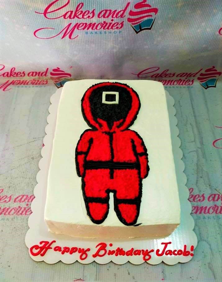 White rectangular cake featuring a large red and black Squid Game guard character piped in icing with a square symbol on the mask. White rectangular cake featuring a large red and black Squid Game guard character piped in icing with a square symbol on the mask.