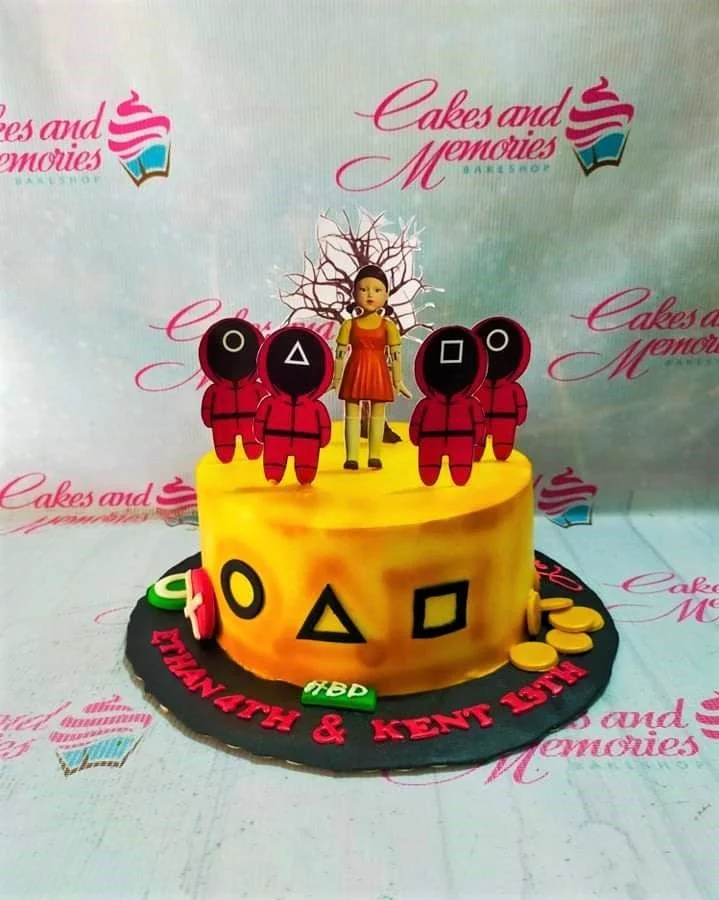 Squid Game themed yellow cake with red guard printouts and a doll figure on top, featuring black geometric symbols on the side.
